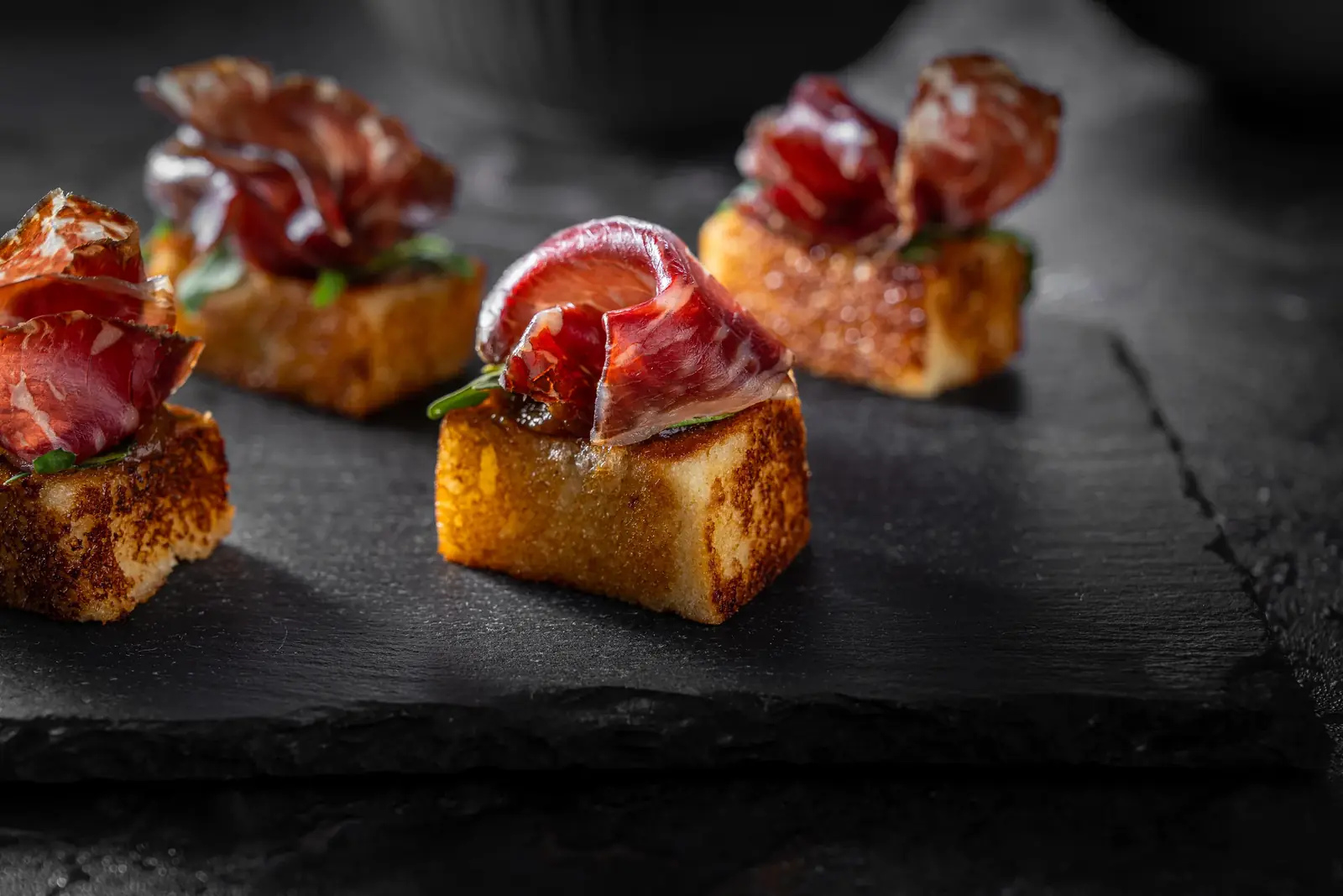 Toasted bread cubes topped with thin slices of cured meat on black slate, catering in Cape Town by myCHEF Cape Town
