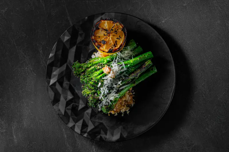 Charred broccolini with grated cheese and a grilled lemon half on a black plate by myCHEF Cape Town